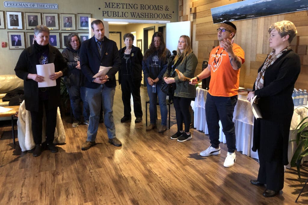 Image from BC Housing of the Second Stage Housing announcement at City Hall. Various people are standing in a circle in the main lobby. Wameesh Tseshaht Chief Councillor Ken Watts delivers a prayer. He is wearing an orange shirt.